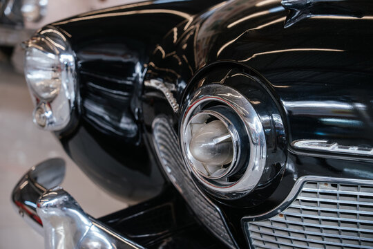 Detailed low-angle view highlights front right fender, headlight, and grille of vintage black automobile. Glossy paint reflects light, showcasing chrome accents and classic design elements.