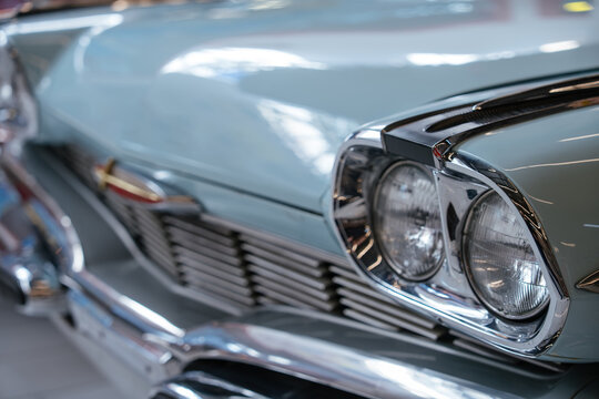 Detailed close-up view of front section of light blue vintage automobile. Polished chrome headlights and grille reflect surroundings, emphasizing classic design in a softly blurred indoor setting.