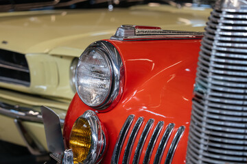 Detailed close-up photograph showcases front-end design of two vintage cars. A vibrant orange car with chrome accents stands out, while a pale yellow car is softly blurred in the background © DenisProduction.com