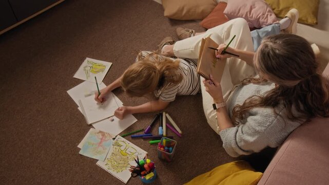 Top view shot of young girl sketching pictures with crayons lying on living room floor while sharing creative hobby with mother