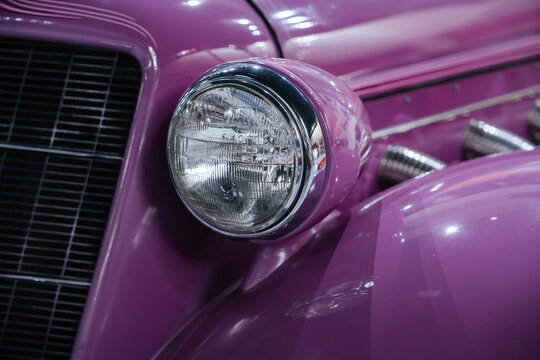 Detailed close-up captures vintage purple automobile's round headlight with chrome rim and textured lens beside a black horizontal-bar grille. Bright lighting enhances glossy finish and chrome exhaust