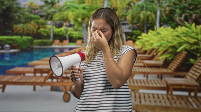 Woman holding megaphone pinches her nose beside a resort swimming pool, eyes closed and wincing while blocking scent with fingers; disgust odor.