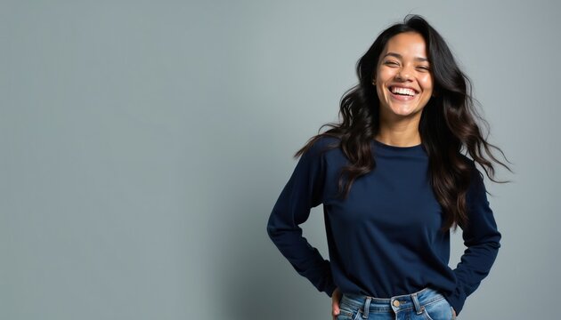 Young woman wears dark blue shirt and jeans. She laughs joyfully with eyes closed, head tilted back. Her dark wavy hair flows freely. Left side offers ample copy space for text.