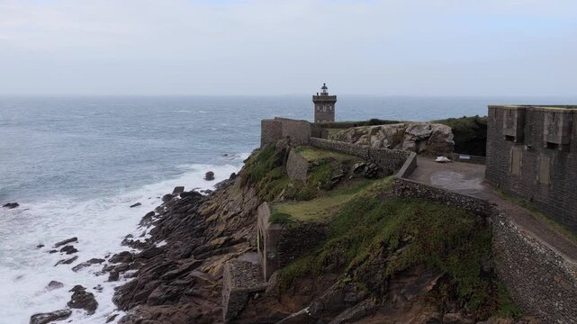 Aerial view of Kermorvan Lighthouse standing on a rocky cliff with stone fortifications and crashing waves under an overcast sky in Le Conquet, Bretagne, France.