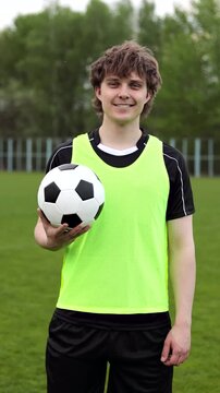 Smiling young man in black football uniform holding a soccer ball on a field. Portrait of an athlete outdoors, concept of sport, fitness, teamwork and healthy lifestyle. Vertical video.