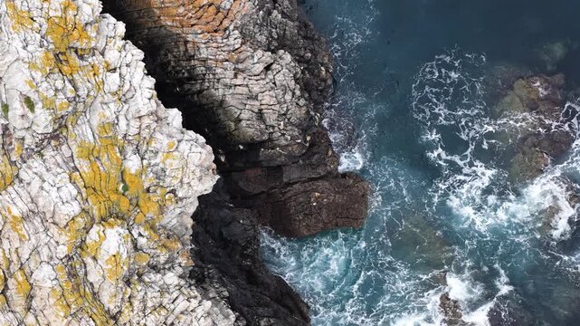 Aerial view of Silence Beach featuring rugged cliffs with yellow lichen and turquoise waves crashing against dark rocks in Asturias, Asturias, Spain.
