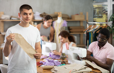 Obraz premium In training tailoring workshop for adult beginners, engaged young guy reviewing paper pattern pinned to fabric surrounded by enthusiastic classmates working on different sewing tasks