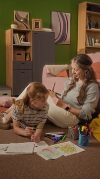 Vertical low eye level shot of girl child lying on floor drawing pictures with colorful crayons then mother showing sketchbook to daughter while spending weekend together in living room