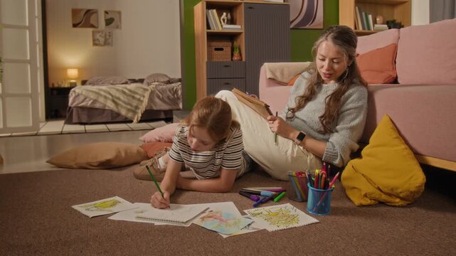 Low eye level shot of preteen girl lying on floor drawing pictures with colorful crayons while chatting with mother during shared activity in living room