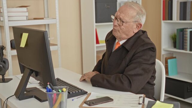 Man clutching lower back at desk in office with computer monitor and paperwork; discomfort aging perseverance.