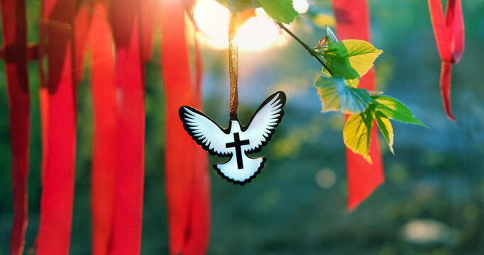 Holy Trinity Sunday, Pentecost day. Dove pendant, red ribbons on tree branch close up, sunny abstract nature background. Christian symbol of Holy Spirit. Christianity religion, faith in God