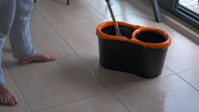 Housekeeper cleaning floor with mop and bucket. Barefoot housekeeper cleaning tiled floor using rotating mop and two-compartment orange-black bucket filled with soapy water