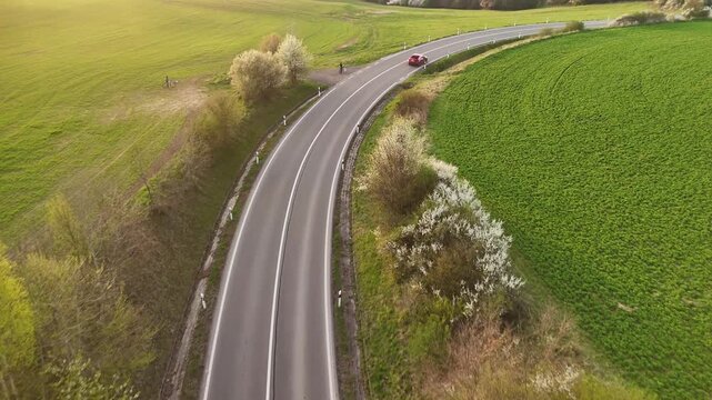 Aerial view of a red car driving on a winding asphalt road through lush green fields and blossoming trees in Cerin, Banska Bystrica Region, Slovakia.