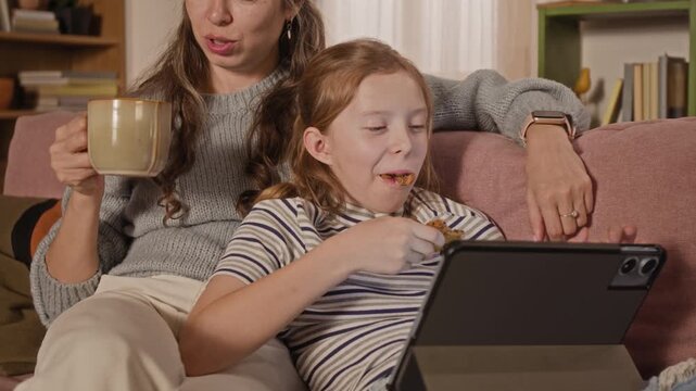 Close up on preteen girl eating biscuit while watching movie on tablet beside mother on living room couch during shared leisure time