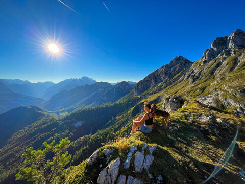 Woman and her mixed breed dog rest at a scenic viewpoint high above the valley at Mangart Pass. Bright summer sun, sweeping ridges, open alpine space and a peaceful mountain moment in the Julian Alps.