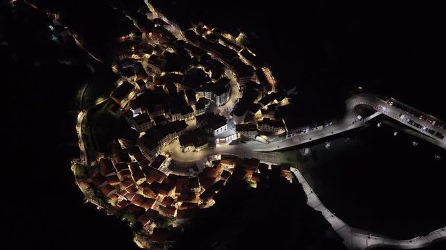 Aerial view of the historic town of Cudillero at night with glowing street lights, traditional houses, and the dark harbor area Cudillero, Asturias, Spain.