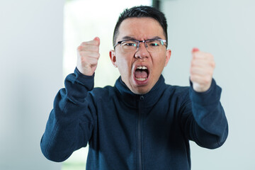 A young Asian man shouts and raises both fists in rage toward the camera, ideal for anger, frustration, or emotional outburst concepts. © fotolite
