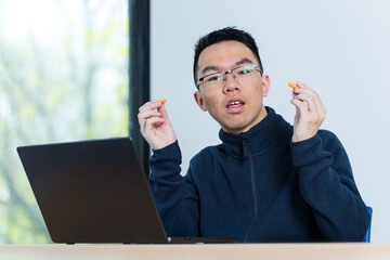 A young Asian male student holds up a pair of orange earplugs at his desk with a laptop open, ideal for neurodiversity, sensory sensitivity, or study productivity concepts. © fotolite