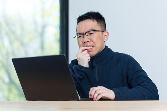 A young Asian male student bites his nails with a worried expression while looking at his laptop, ideal for anxiety, ADHD, or academic stress concepts.