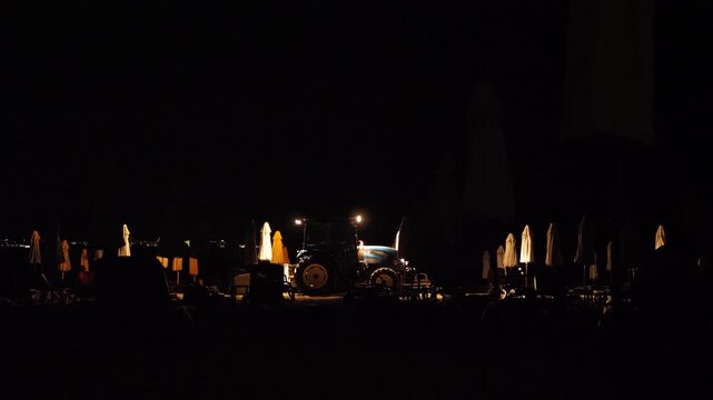 Night seaside scene, sand sifting tractor night work. Serene nighttime shoreline with tractor grooming among umbrellas. Nighttime coastal scene with maintenance activity and subdued lighting