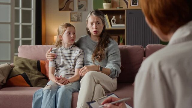 Adult mother checking young daughters temperature by touching girls forehead while counselling female pediatrician about symptoms during home visit