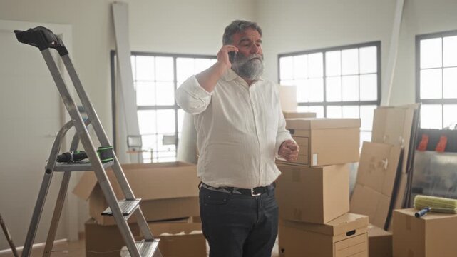 Man holds smartphone to ear among stacked moving boxes and ladder in a sunlit building; moving planning concern.