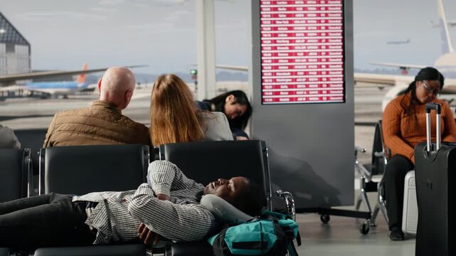 Multiethnic passengers in airport resting on bench after cancelled flight shown on flight status display. African american people sleeping on chairs after losing flight due to airline overbooking