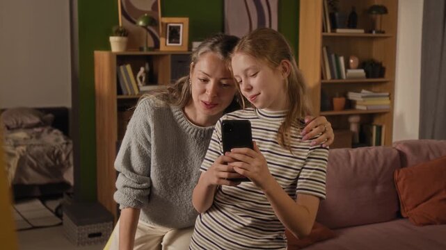 Medium full shot of cheerful mother and young daughter pulling funny faces while taking selfie on smartphone looking in mirror at home