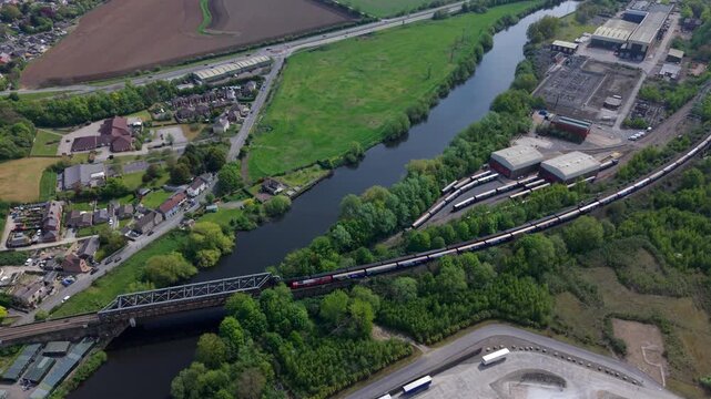 Aerial view of a long freight train crossing Ferrybridge over the River Aire with industrial buildings and green fields Knottingley, England, United Kingdom.