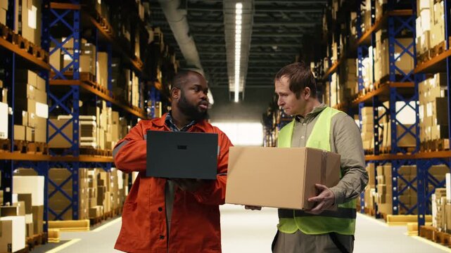 Diverse colleagues moving cargo in boxes using laptop for inventory control in large business warehouse. Distribution hub staff coordinate order fulfillment and shipping operations.