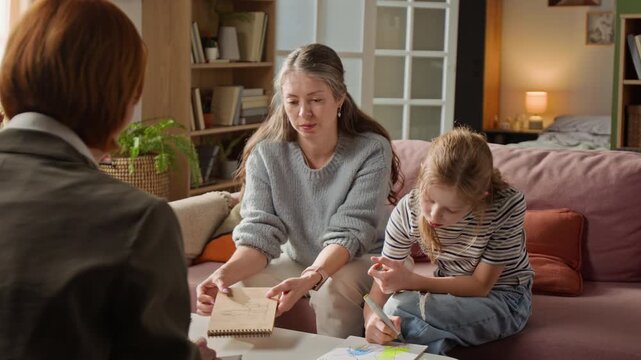 Adult mother and young daughter sitting on living room couch showing colorful drawings to therapist while specialist assessing family through creative task during home based consultation