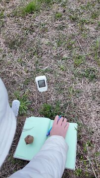 A close-up field setup shows soil pH and temperature meters inserted into the ground while a hand records measurements in a notebook placed on the soil. The scene captures practical environmental data