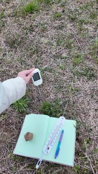 A hand places and uses soil testing instruments in an outdoor field setup, including a pH meter and soil thermometer, while recording observations in a notebook stabilized with a stone. The scene