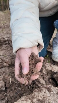 Low-angle close-up shot of legs approaching the camera, followed by a crouching motion where a hand picks up a clump of soil and brings it toward the lens. The soil is squeezed and crumbles, falling