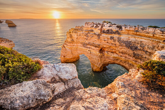 Sunrise view of Praia da Marinha beach in Algarve, Portugal, with a limestone sea arch, Atlantic water, warm sky, and rugged coastal cliffs.