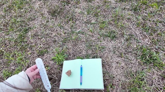 A hand conducts soil field measurements by inserting a soil thermometer followed by a pH meter into the ground while recording results in a notebook placed on the soil and stabilized with a stone. The