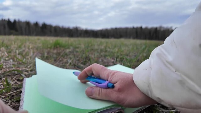 Close-up ground-level scene of dry grass with small green sprouts where a hand places an open notebook and holds a pen. Pages move in the wind and are stabilized before writing begins. Suitable for