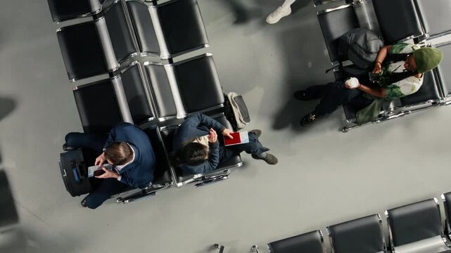 Walking, devices and travelers in airport terminal next to people on chairs. Waiting for plane, trolleys and passengers with trolleys navigating air transportation hub, top down view shot
