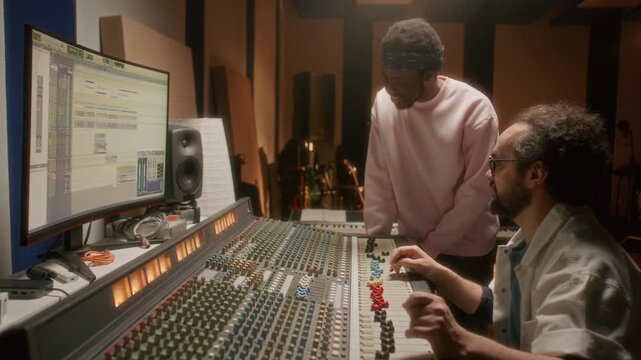Professional sound engineer adjusting faders on mixing console and discussing audio tracks with young African American rapper while working together in music recording studio