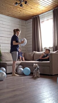 Girl juggling balloon while dog watches, brother reaches from sofa and giggles as silver and blue balloons scatter across hardwood, lively household moment with warm ceiling light