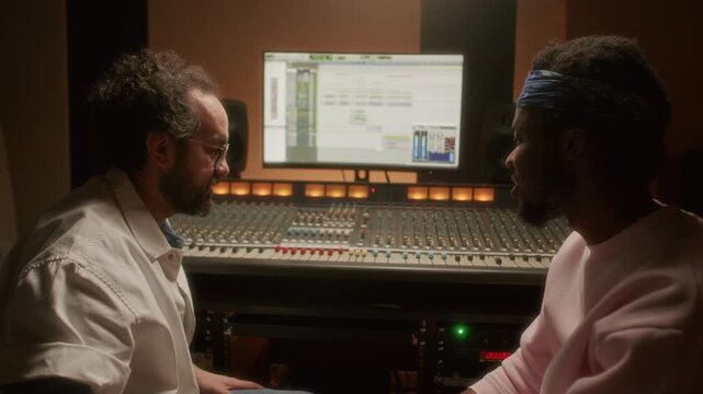 Caucasian audio engineer and African American musician having discussion, operating mixing desk and looking at computer screen with audio editing software during studio recording session