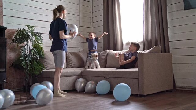 Children rehearsing celebration with blue balloons, older sibling coordinates playful cheer as younger jumps on couch and dog explores, scattered silver balloons and sunlight create messy joyful