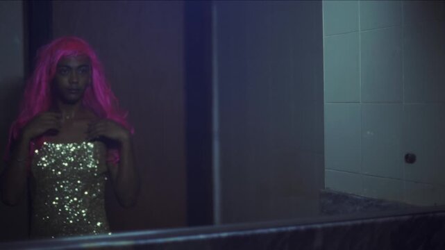 Transgender woman wearing a pink wig and a sequin top looking at her reflection in the mirror