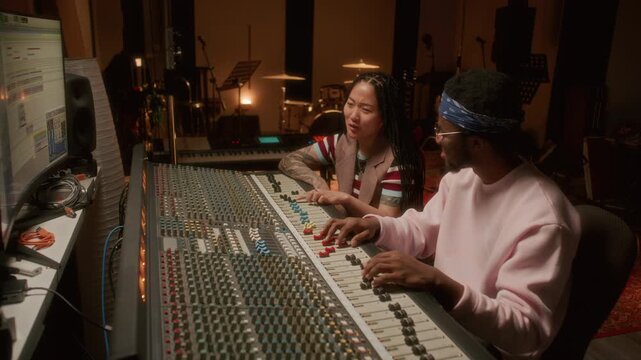 Young Asian female music producer collaborating with African American audio engineer, smiling and speaking while mixing soundtracks in recording studio