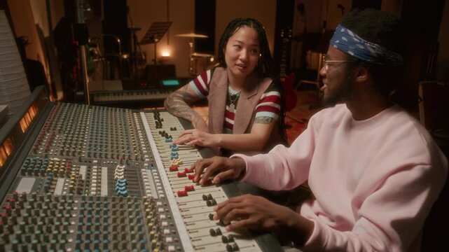 Asian female music producer and African American male audio engineer adjusting sound levels on mixing console, enjoying beat and having discussion during studio recording session