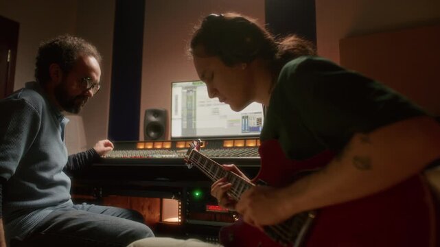 Young guitarist in headphones playing red electric guitar while sound producer monitoring audio tracks on mixing console and digital screen during music recording session in studio