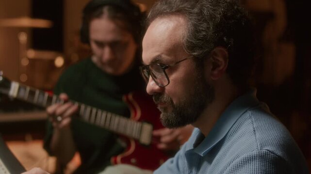 Music producer operating professional audio console while young male guitarist practicing in recording studio with modern equipment. Tilt up shot, rack focus