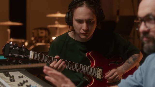 Audio engineer controlling professional mixing console while young music artist playing electric guitar during recording session in studio. Close-up, tilt down shot