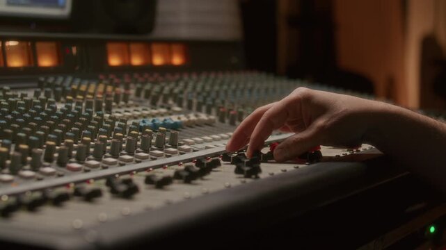 Hand of audio engineer adjusting sliders on professional analog mixing console during studio recording session, rows of colorful knobs and illuminated VU meters visible in background. Close up shot