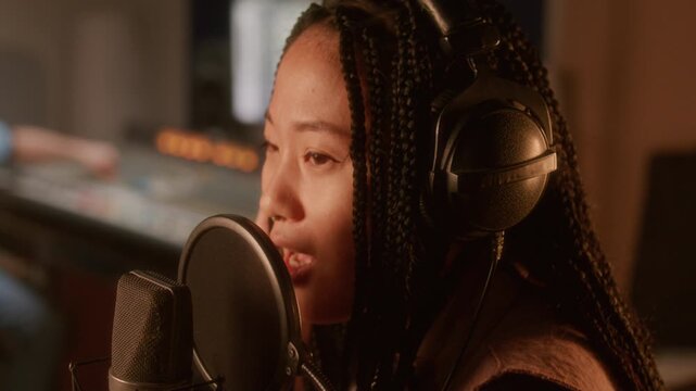 Young Asian female singer with long braids and headphones singing into professional condenser microphone during vocal recording session in music studio. Close up shot